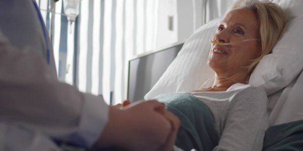 Close up of doctor holding hand and talking to sick aged female patient in hospital ward. Ill senior woman with nasal oxygen tube lying in bed and talking to doctor